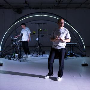 Ein Performer in weißem Shirt steht vor einem transparenten Kuppelzelt und spricht. Hinten ist eine weitere Person in weißem Hemd und bedient Technik.