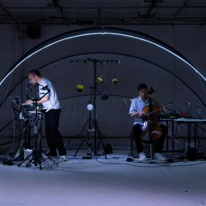 Zwei Performer befinden sich im offenen transparenten Kuppelzelt. Die Person rechts spielt Cello, die andere steht über ein Laptop gebeugt. 