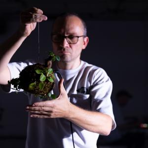 Ein Performer im weißen Hemd hält eine Kugel aus Moos mit kleinen Pflanzen darauf in den Händen. In der Kugel steckt ein Kabel.