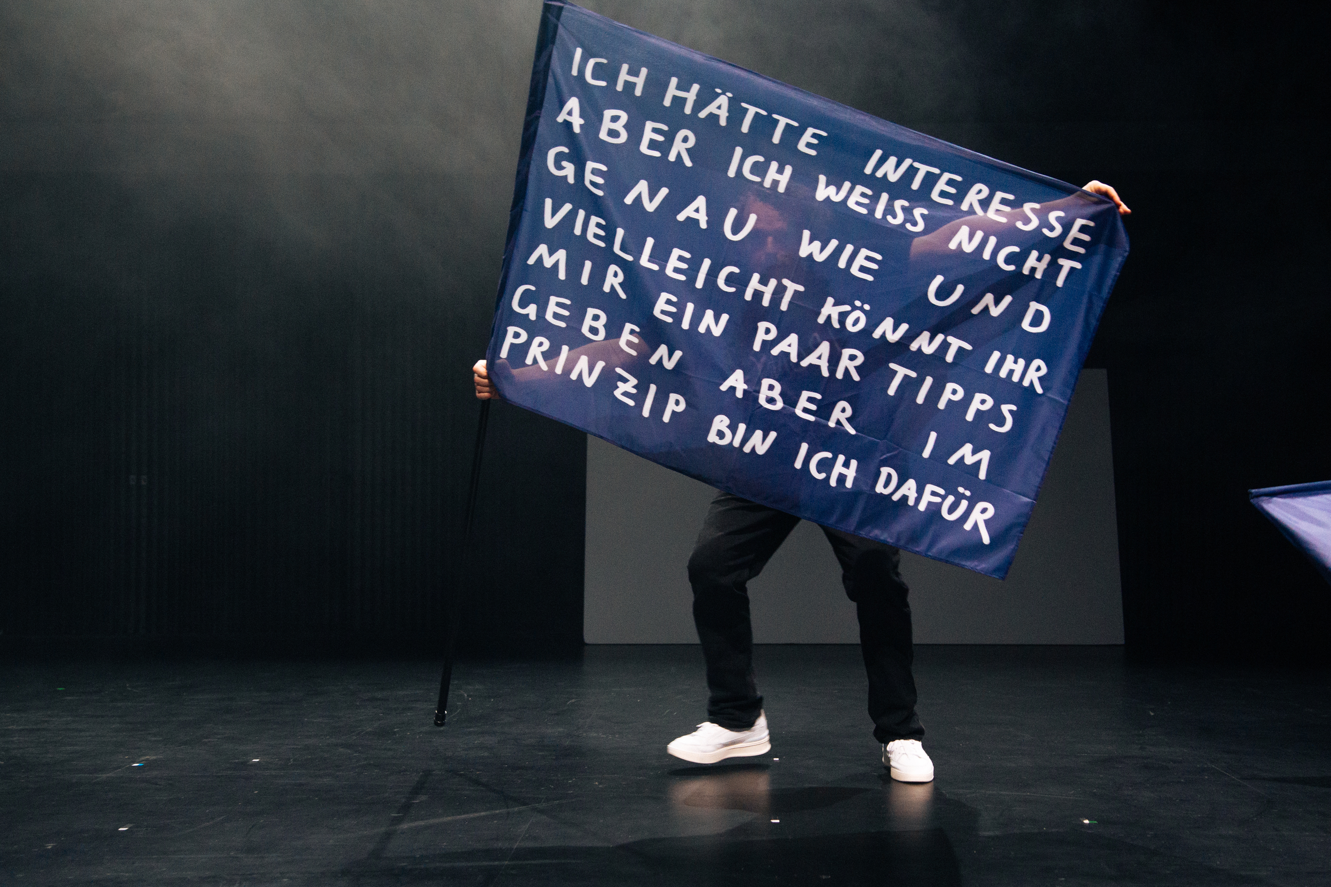 Eine Person steht auf einer dunklen Bühne und hält eine große blaue Fahne an einem schwarzen Stab. Die Fahne trägt in weißer Handschrift den Text: „Ich hätte Interesse aber ich weiss nicht genau wie und vielleicht könnt ihr mir ein paar Tipps geben aber im Prinzip bin ich dafür." Der Oberkörper der Person ist hinter der Fahne verborgen, sichtbar sind nur Beine in dunkler Hose und weiße Sneaker. Der Bühnenhintergrund ist dunkel mit leichtem Nebel.